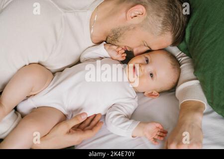 Ein hübscher, junger Vater liegt im Bett im Schlafzimmer und umarmt seinen liebend lachenden sechs Monate alten Jungen. Das Konzept der bedingungslosen Liebe und Vaterschaft Stockfoto