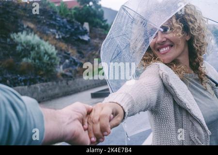 Pov eines Mannes, der die Hand einer Frau unter dem Regen hält. Ein glückliches junges erwachsenes Paar genießt an regnerischen Tagen Freizeitaktivitäten im Freien mit Regenschirm und lächelndem eac Stockfoto