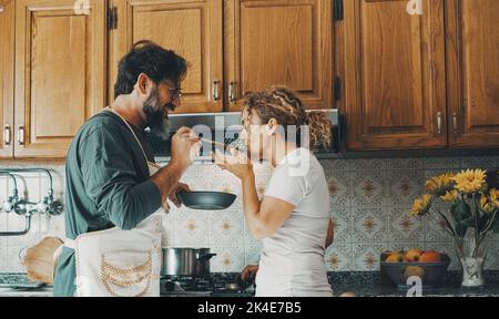 Ein glückliches echtes Paar zu Hause, das das Leben zusammen und die Beziehung genießt. Mann, der der Frau Essen zum Geschmack gibt. Männliche Leute, die für seine Frau kochen. Männlich und f Stockfoto