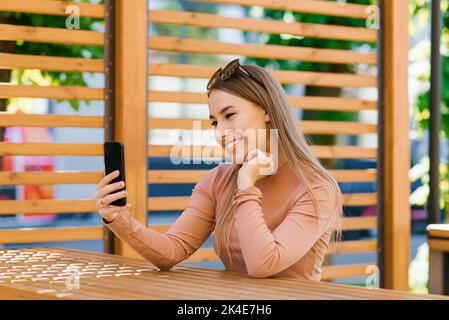 Junge hübsche Bloggerin spricht per Videolink oder Übersetzung in einem Café im Freien Stockfoto