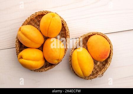 Mehrere reife gelbe Ananas-Aprikosen in Strohtellern, Nahaufnahme, auf einem weißen Holztisch, Draufsicht. Stockfoto