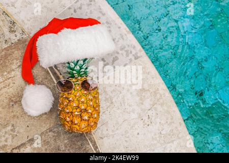 Blick von oben auf lustige Ananas in Sonnenbrillen und Weihnachtsmannshut, die bei sonnigem Wetter in den Tropen am Rand des Pools liegt und sich sonnt. Weihnachten in Stockfoto