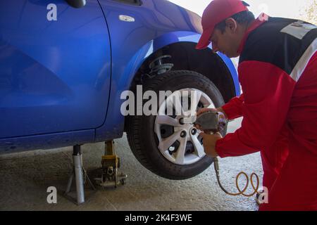 Bild eines Mechanikers in seiner Werkstatt, der mit einem pneumatischen Schraubendreher das Rad eines auf Böcken ruhenden Autos zerlegt. Reparaturarbeiten für das Auto Stockfoto