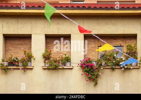 Urlaub in einer alten europäischen Stadt. Fenster eines alten Wohnhauses mit Holzjalousien und roten Blumen, festlichen bunten Fahnen Stockfoto