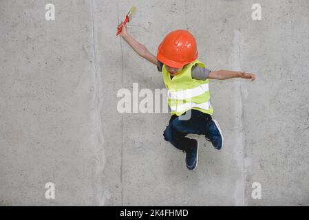 Junge mit Hardhut und Sicherheitsweste, der vor einer Betonwand springt Stockfoto