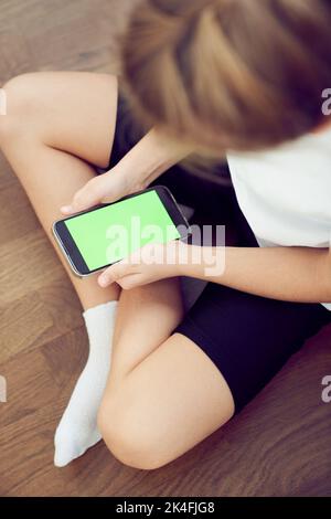 Hände von Kindern mit einem grünen Bildschirm. Ein kleines Mädchen sitzt auf dem Boden und hält ein Smartphone mit einer Chromakey in den Händen. Blick aus einem großen Winkel. Hochwertige Fotos Stockfoto