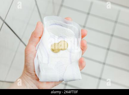 Hygiene Armut Konzept, junge Frau hält einmal Menstruationsauflage und Münzen in der Hand in Hause Badezimmer. Kann sich keine neuen leisten. Stockfoto