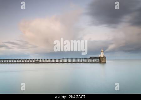Eine lange Belichtung bei Dämmerung an einem kalten Tag im Blyth Harbour an der Küste von Northumberland, mit einem hölzernen Pier und einem kleinen weißen Leuchtturm Stockfoto