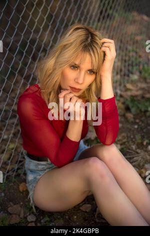 Hoher Winkel von wunderschönen jungen weiblichen Modell in Mini-Denim-Shorts und rote Bluse berühren lange blonde Haare und Blick weg in der Nähe Kettengliedzaun in Zählung Stockfoto