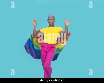 Afroamerikanischer Mann in farbenfroher Kleidung mit Make-up, der auf die Kamera schaut und hinter dem Rücken eine Regenbogenfahne ablässt, während er gegen türkisfarbene Backgroun tanzt Stockfoto