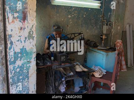 Ein Mann mittleren Alters mit Mütze sitzt auf einer alten, antiquierten Singer-Nähmaschine und arbeitet in einer Fabrik in Havanna, Kuba. Stockfoto