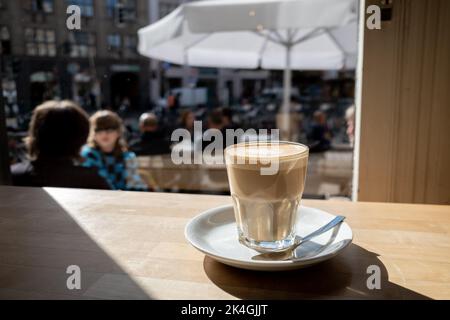 Flacher weißer Kaffee aus transparentem Glas und weiße Teller lagen auf einer rauen Holztischtheke neben den Fenstern des Cafés. Stockfoto