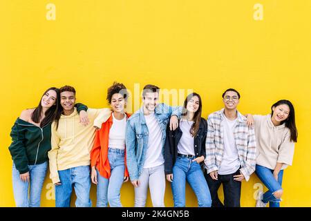 Lächelnde junge Gruppe von multirassischen glücklichen Freunden, die an der gelben Wand gelehnt sind Stockfoto