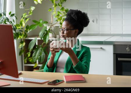 Erfolgreiche nachdenkliche afroamerikanische Frau, die Kaffee trinkt, sitzt am Tisch mit Computer im Heimbüro Stockfoto
