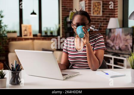 Fernstudentin, die sich zu Hause eine Lehranleitung auf dem Laptop ansieht und Kaffee trinkt. Frau, die an Online-Kursen teilnimmt, junge Freiberuflerin, die im Internet arbeitet, hält einen Teebecher Stockfoto