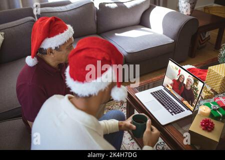 Biracial Vater und erwachsenen Sohn in santa Hüte machen Laptop weihnachten Videoanruf mit lächelndem Paar. weihnachten, Festlichkeit und Kommunikationstechnik. Stockfoto