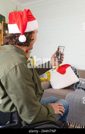 Kaukasischer Mann im Rollstuhl und weihnachtsmann Hut macht weihnachten Smartphone Video-Anruf mit männlichen Freund. weihnachten, Festlichkeit und Kommunikationstechnik.