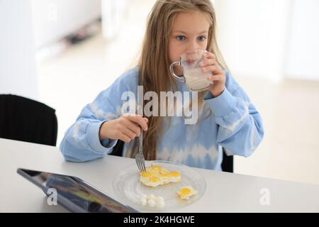 Das kleine Mädchen trinkt morgens Milch mit Ei und sitzt am Tisch. Richtige Ernährung für Kinder Stockfoto