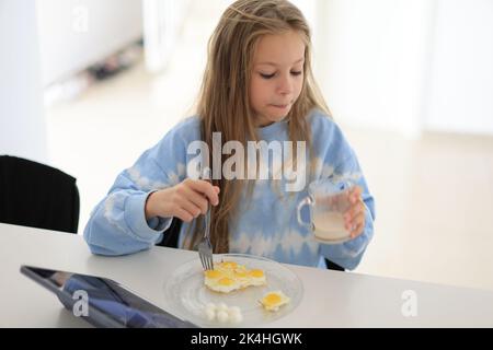 Das kleine Mädchen trinkt am Morgen die Milch der ganzen Kuh mit dem Ei und sitzt am Tisch. Richtige Ernährung für Kinder Stockfoto