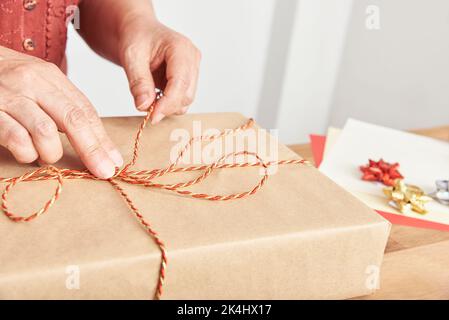Hände einer unbekannten reifen Frau, die ein Weihnachtsgeschenk mit einer dekorativen Schnur bindet. Helles Design mit Platz für Kopien. Konzept der Verpackung von Urlaubsgeschenken. Stockfoto