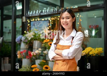 Attraktive junge asiatische Floristin, Arme überkreuzt lächelnd und schauend auf die Kamera, stehend vor ihrem Blumenladen. Small Business Concept Stockfoto