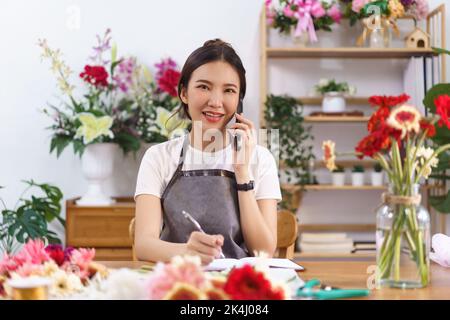 Floristen-Konzept, weibliche Floristin im Gespräch mit dem Kunden am Telefon und Notizen im Notebook im Geschäft. Stockfoto