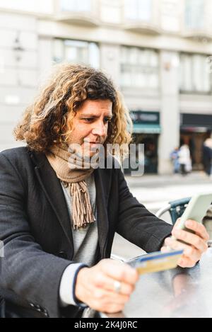 Ein männlicher Kunde mittleren Alters in eleganter Freizeitkleidung mit Kreditkarte, der sein Mobiltelefon zum Online-Shopping verwendet, während er am Tisch im Straßencafé sitzt Stockfoto