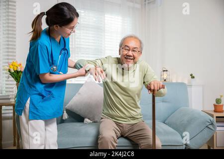 Internationaler Tag für ältere Menschen. Junge Pflegerin unterstützt den älteren Mann beim Aufstehen mit Gehstock vom Sofa, Krankenschwester trägt blaue Uniform und hilft ihm dabei Stockfoto