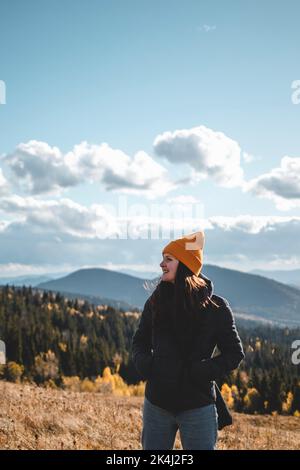 Junge glückliche Frau in oranger Mütze steht vor der Berglandschaft Stockfoto