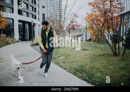 Glücklicher junger Mann, der während des Herbsttages seinen Hund im Freien in der Stadt herumläuft. Stockfoto