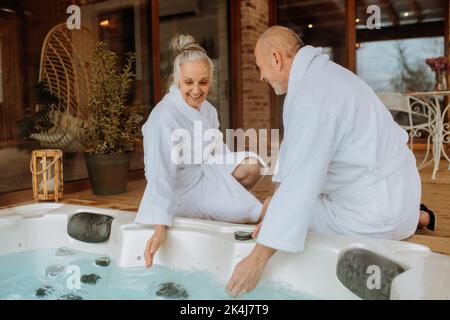 Das ältere Paar im Bademantel überprüft die Temperatur im Whirlpool im Freien und bereitet sich auf das Baden mit seiner Frau vor. Stockfoto