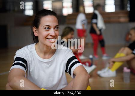 Eine Gruppe von jungen und alten Frauen, Sportmannschaftsspielern im Fitnessstudio, die nach dem Spiel sitzen und sich ausruhen. Stockfoto