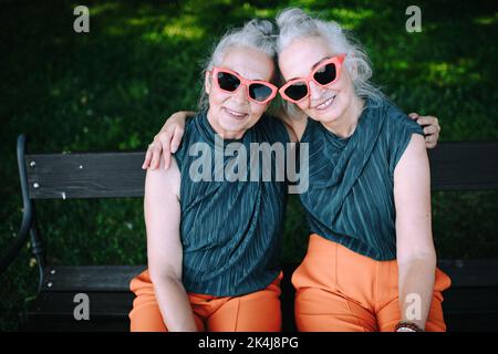Blick aus der Höhe auf glückliche ältere Frauen, Zwillinge in derselben Kleidung, lächelnd und posiert im Stadtpark. Stockfoto