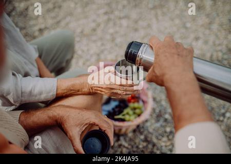 Nahaufnahme eines älteren Paares, das am Herbsttag am Strand picknickt und Tee aus der Thermoskanne trinkt. Stockfoto