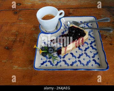 Espresso-Kaffee in einer weißen Tasse mit Cheesecake Pie Scheibe mit Heidelbeerfrucht und Sirup mit Löffel und Gabel auf einem Teller, Süßes Essen gekrönt Stockfoto