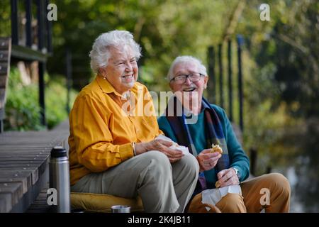 Glückliches Seniorenpaar, das Picknick macht und sich nach dem Spaziergang in der Nähe des Sees ausruhen kann. Stockfoto