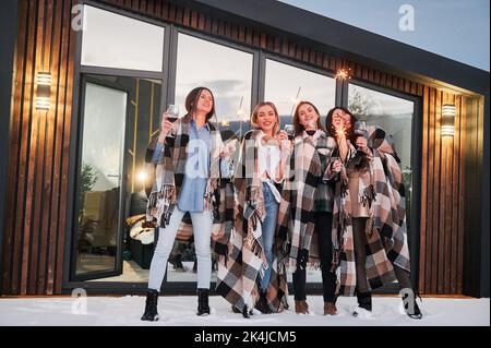 Junge Frauen genießen die Winterwochenenden auf der Terrasse des modernen Barnhouse. Vier Mädchen in Plaids trinken Wein und feiern mit Wunderkerzen am Abend. Stockfoto