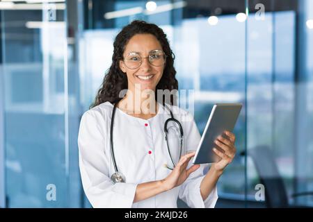 Porträt einer lateinamerikanischen Ärztin. Im Flur des Krankenhauses stehend. Er arbeitet, hält die Tablette in den Händen, benutzt sie, typiert. Er schaut lächelnd auf die Kamera. Stockfoto