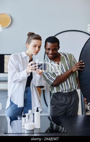 Vertikales Porträt zweier junger Fotografen, die im Studio an Produktfotografie arbeiten und Mobiltelefone für soziale Medien nutzen Stockfoto