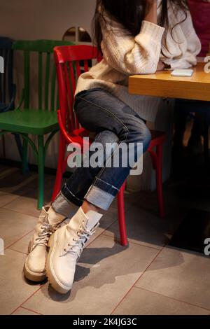 Nahaufnahme von Details der lässigen Damenmode. Beine aus Denim-Hose und hohe Leder-Stiefeletten mit Schnürsenkeln. Frau sitzt am Tisch im Café Stockfoto