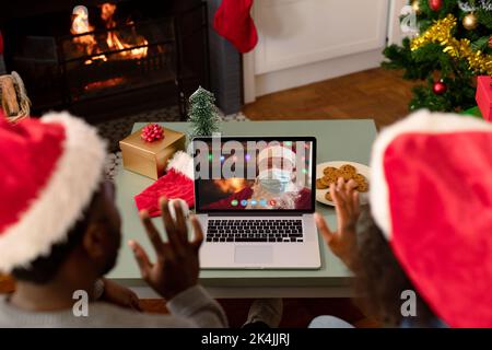 Winkende afroamerikanische Paar macht Laptop weihnachten Videoanruf mit dem weihnachtsmann in Gesichtsmaske. weihnachten, Festlichkeit und Kommunikationstechnik Dur Stockfoto