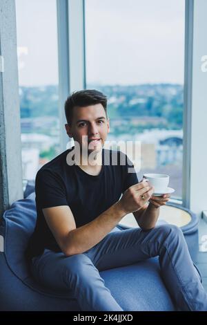 Ein junger Mann sitzt auf einem Stuhl mit Sitzsack in der Nähe eines großen Fensters im Büro und trinkt während seiner Mittagspause Kaffee. Ein Mann in einem schwarzen T-Shirt und einer Hose Stockfoto