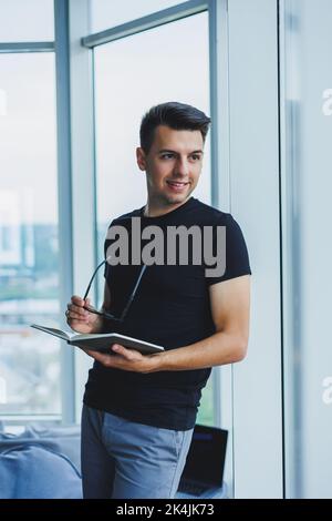 Ein studentischer Manager steht in der Nähe eines großen Fensters und liest Notizen in einem Notizbuch. Ein junger Mann in einem schwarzen T-Shirt mit Brille steht im Büro in der Nähe des Stockfoto