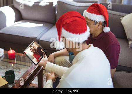 Biracial Vater und erwachsenen Sohn in santa Hüte machen Tablet weihnachten Videoanruf mit Paar. weihnachten, Festlichkeit und Kommunikationstechnik. Stockfoto