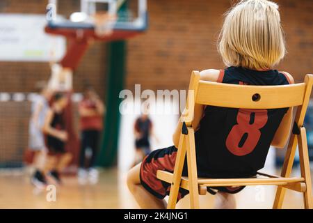 Ein junger Basketballjunge sitzt auf einer Ersatzbank und wartet darauf, ins Spiel einzutreten. Basketball-Trainingskurs für Kinder Stockfoto