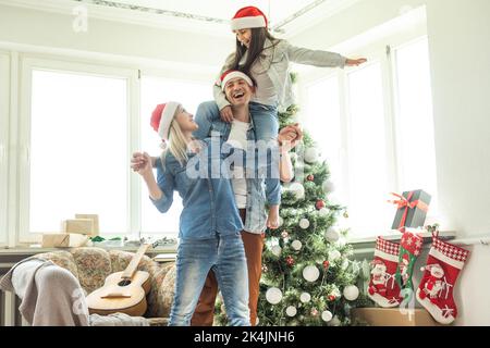 Familie schmücken einen Weihnachtsbaum. Junger Mann mit seiner Tochter auf seinen Schultern hilft ihr, den Weihnachtsbaum schmücken. Stockfoto