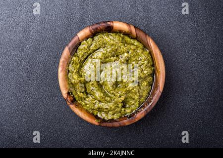 Draufsicht auf Pesto-Sauce in Holzschüssel als Zutat für leckere Pasta-Gerichte auf schwarzem Beton-Hintergrund Stockfoto