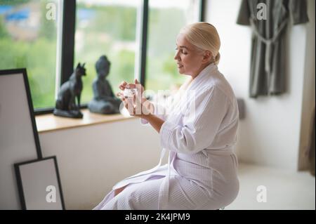 Reife Frau im Frottee-Bademantel ein Hautpflegeprodukt auftragen Stockfoto