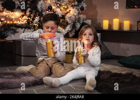 Entzückende Kinder, Junge und Mädchen halten gelbe Kerzen und machen Wunsch. Bruder und nette Schwester sitzen auf dem Boden in dunklen Raum. Weihnachtsbaum und Präs Stockfoto