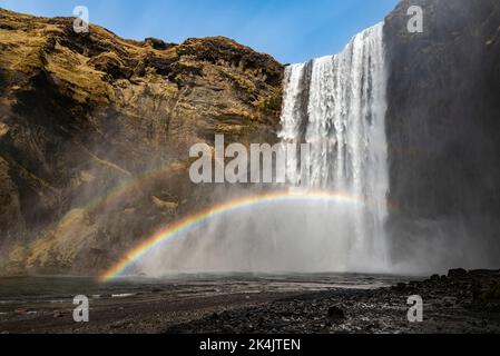 Niedriger Winkel des mächtigen Skógafoss-Wasserfalls unter einem klaren blauen Himmel, mit einem schönen doppelten Regenbogen, in der Nähe der Route 1 / Ring Road, Island Stockfoto